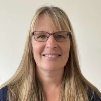 Smiling woman with long blonde hair and glasses, wearing a necklace, against a plain light background.