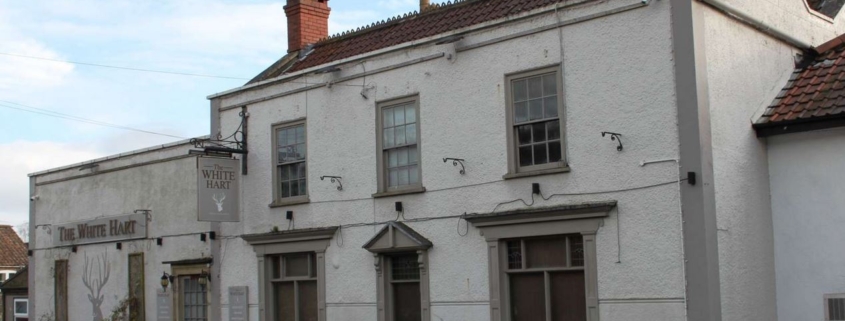 White-painted building with sign reading "The White Hart," featuring multiple windows and potted plants outside.