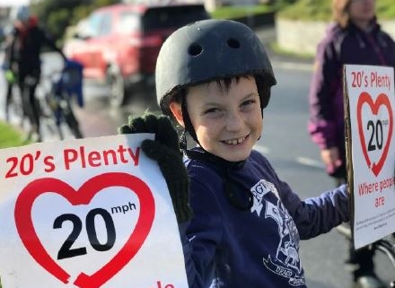 Smiling child in a helmet holds “20’s Plenty” speed limit signs during a street safety event.