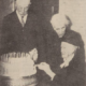 Three elderly people stand around a large cake with many candles, one person is about to cut the cake.