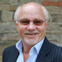 Smiling older man with glasses and a goatee, wearing a pinstripe suit, standing by a stone wall.